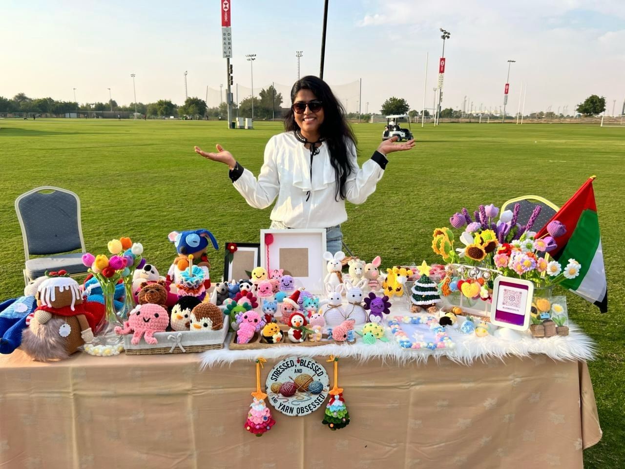Shilpa smiling at her Loomé market stall on a grass field, with the UAE flag and colourful crochet flowers on the table.